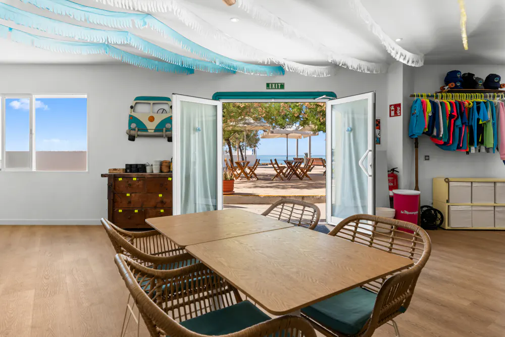 Interior view of a room with a wooden table and chairs, leading to a beach view through open doors. Blue and white decorations hang from the ceiling.