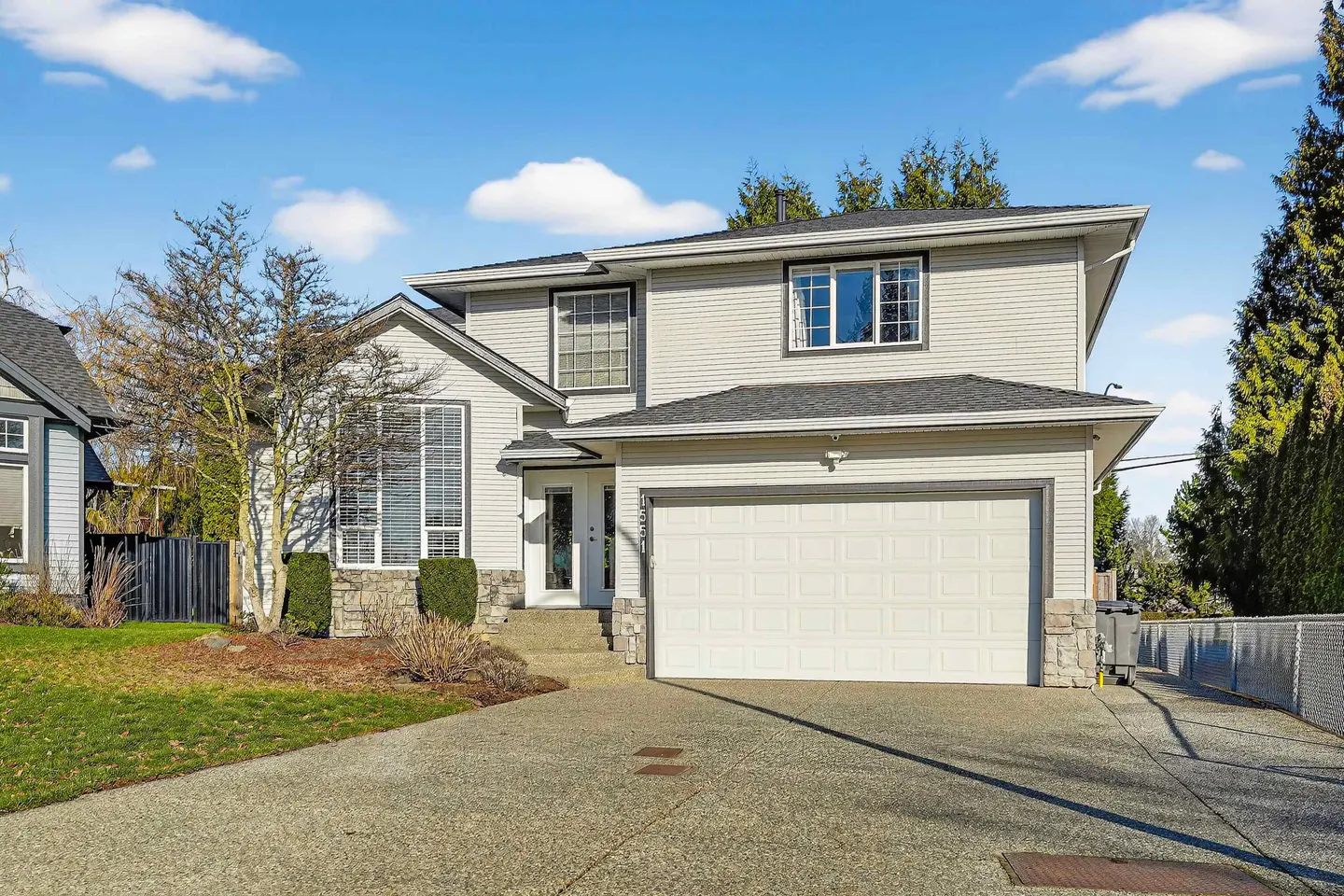 Two-story house with light gray siding, a white garage door, and a concrete driveway on a sunny day.