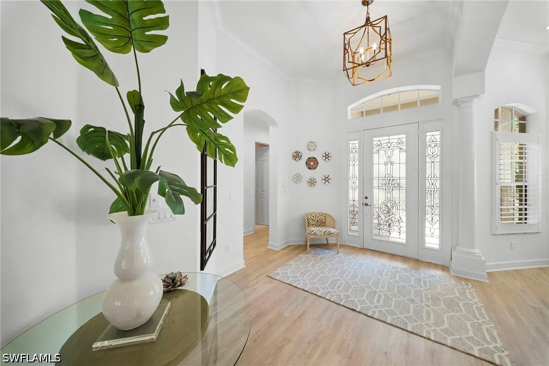 Bright foyer with white walls, light wood floors, and a geometric rug. A monstera plant sits on a glass table. Decorative snowflakes adorn the wall.