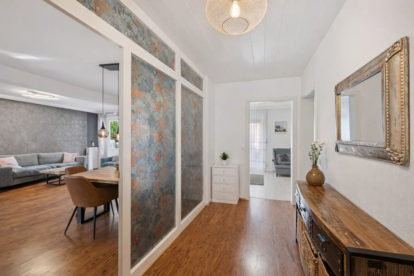 A bright hallway with wood floors, a console table, and a framed mirror. A living room with a gray sofa is visible through a decorative partition.