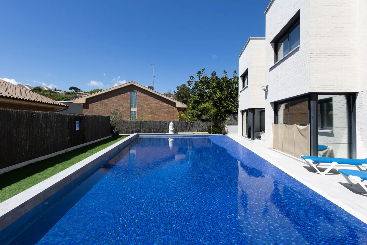 A bright blue tiled pool sits beside a modern white house under a clear blue sky. Lounge chairs are visible near the house.