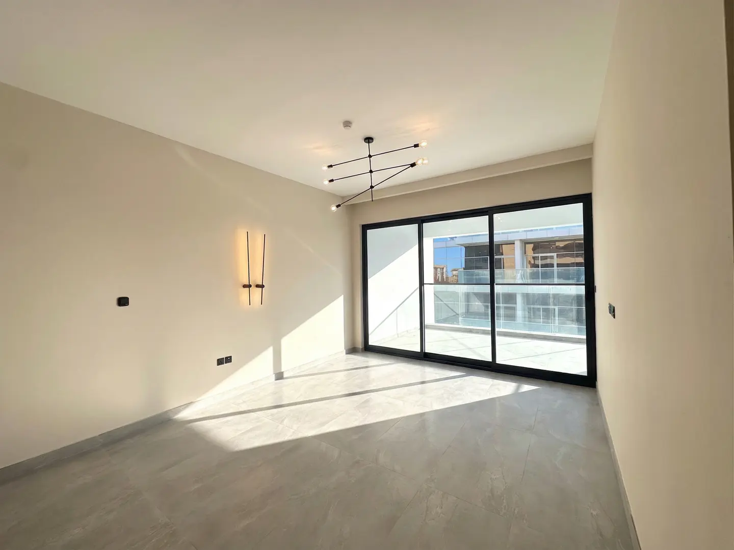 Empty room with gray tile floor, beige walls, and black framed sliding glass doors to a balcony. Modern light fixture on the ceiling.