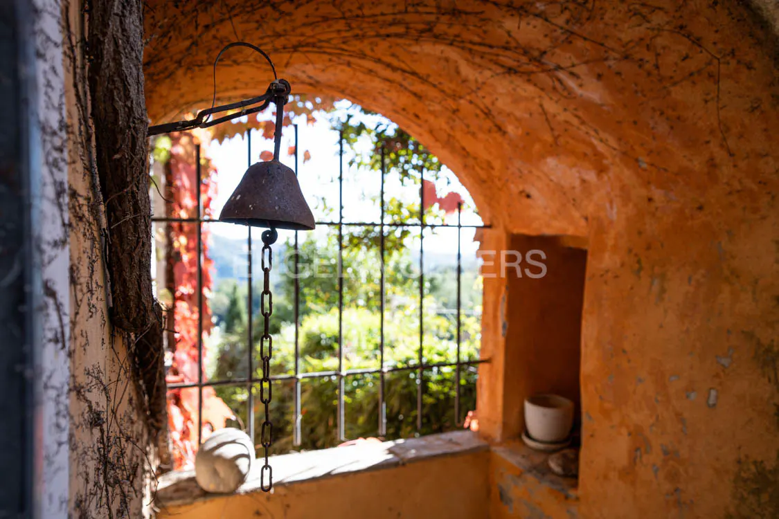 Arched window view with a hanging metal bell. The orange wall is textured, with vines. Outside, trees and a distant landscape are visible.