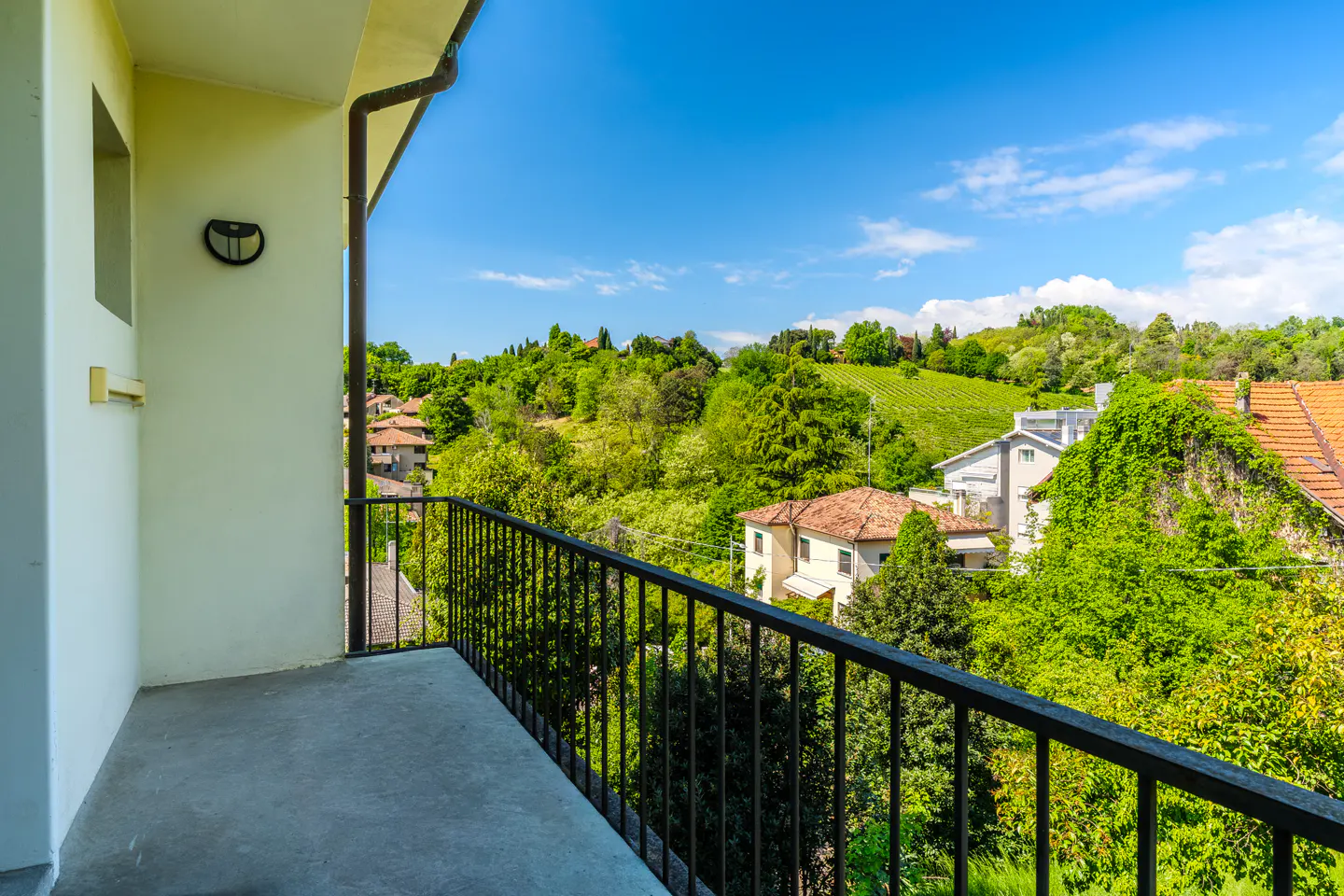 Balcony view with black railing overlooking lush green hills, houses, and a vineyard under a blue sky.