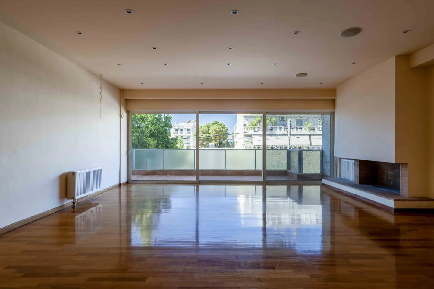 An empty room with hardwood floors, white walls, and a large window with a view of trees and buildings. A fireplace is on the right.