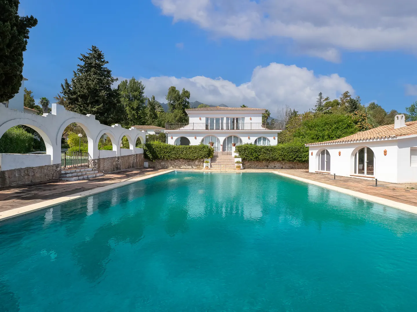 A large turquoise pool in front of a white two-story house with arched walkways and green trees under a blue sky.