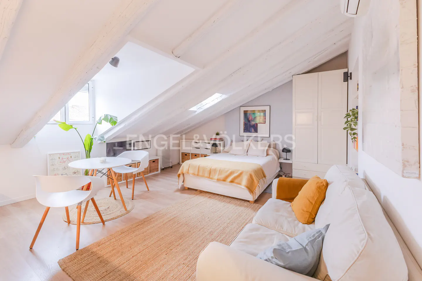 Bright attic apartment with white walls, exposed beams, and skylight. Features a bed, sofa, dining table, and wardrobe.