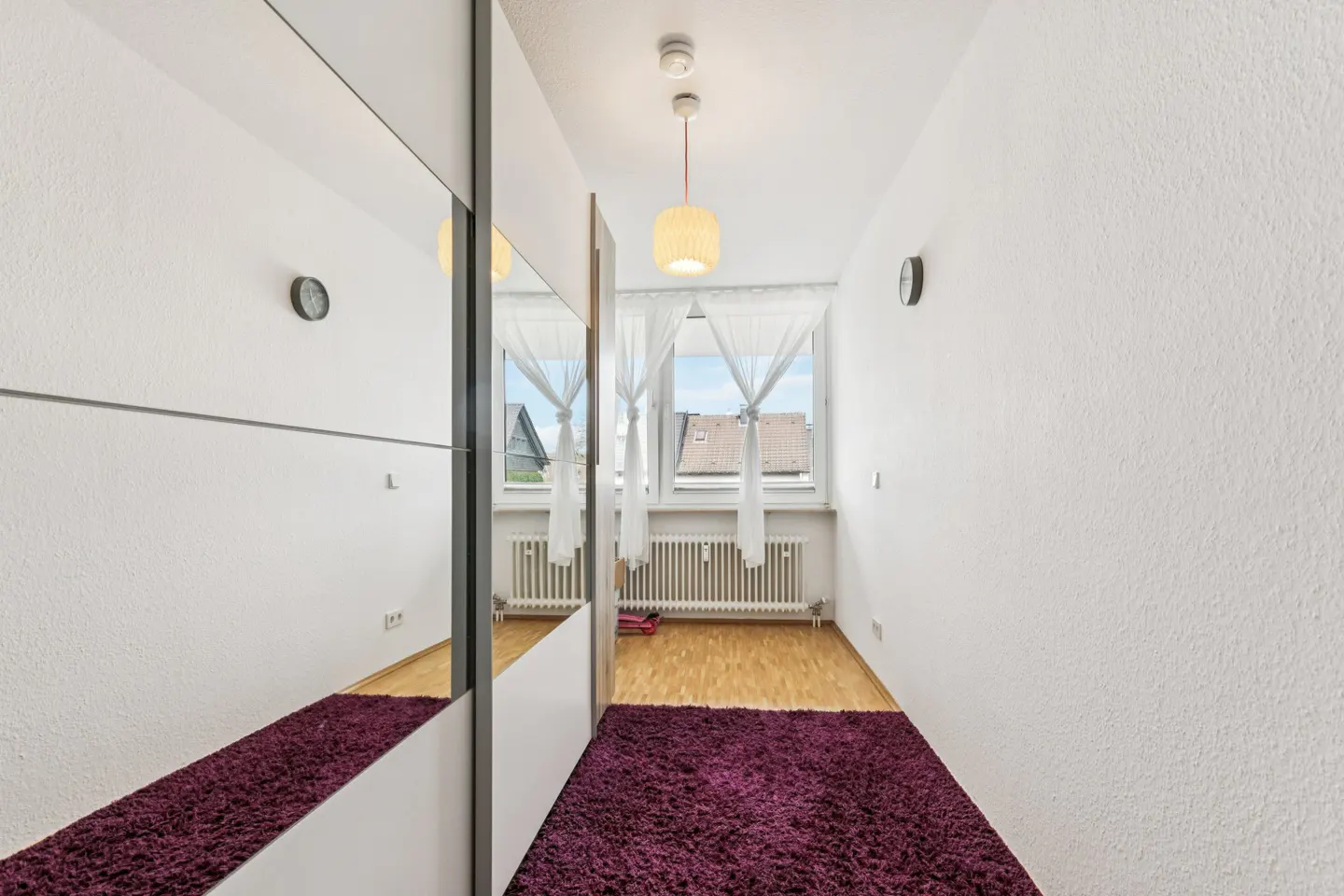 Hallway with a purple rug, wood floor, and white walls. A mirrored closet is on the left, and a window with white curtains is at the end.