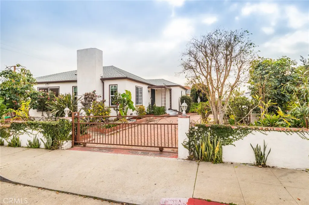 A white single-story house with a brown gate and lush landscaping.