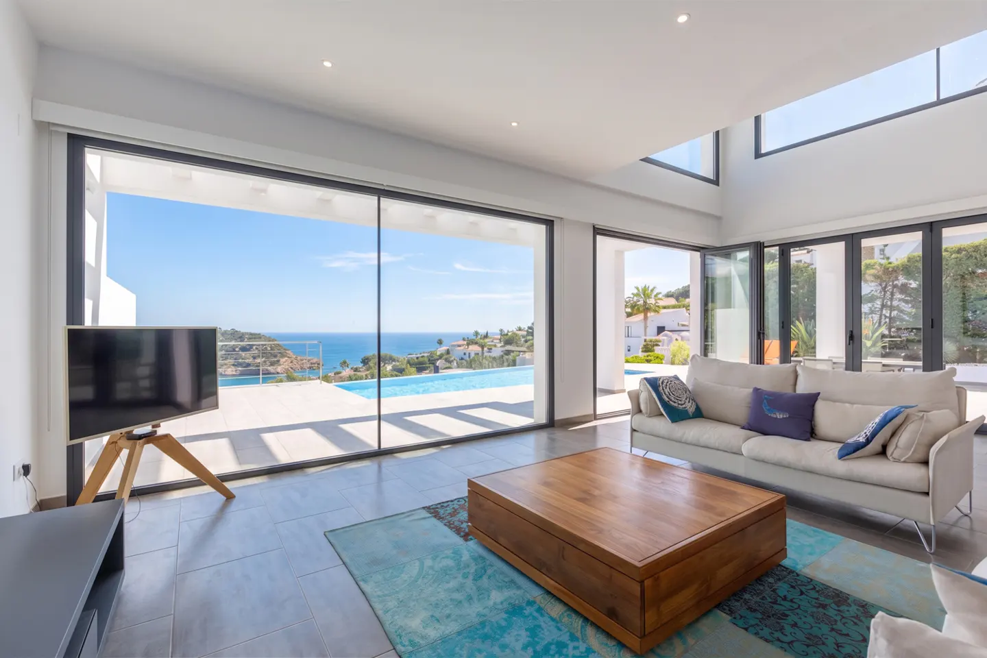 Bright living room with a large window view of the ocean and pool. A beige sofa, wood table, and TV are inside.