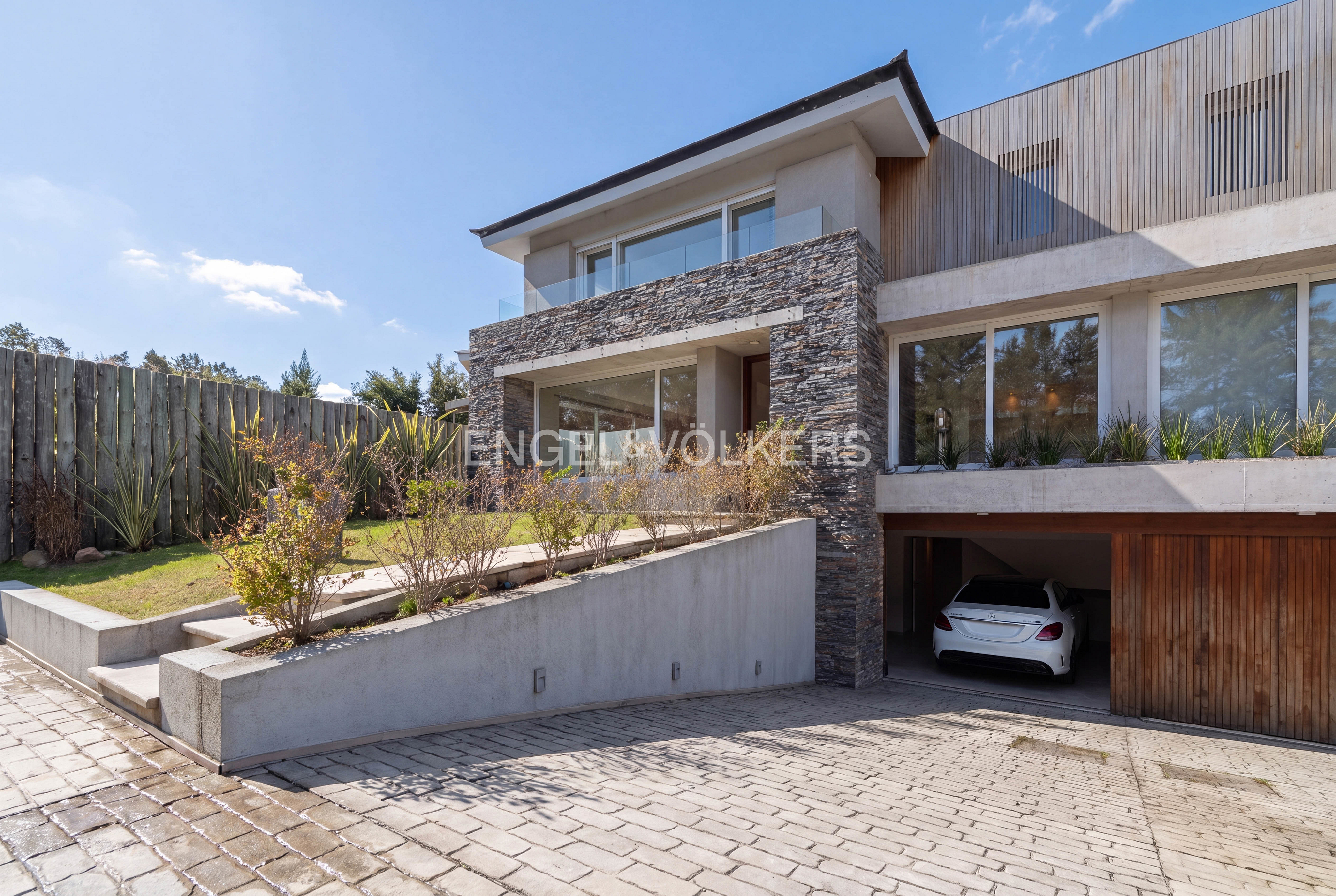 Modern two-story home with stone facade, concrete retaining wall, wooden garage door, and a white car parked in the driveway.