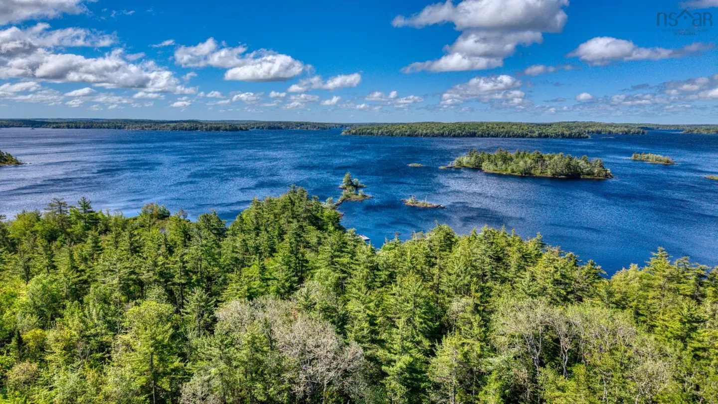 Aerial view of a lush green forest overlooking a deep blue lake with small islands under a bright, cloudy sky.