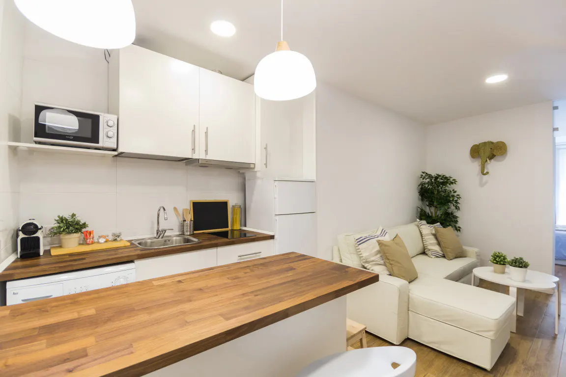 Bright studio apartment with white cabinets, wood countertops, and a white sofa. An elephant head decorates the wall.