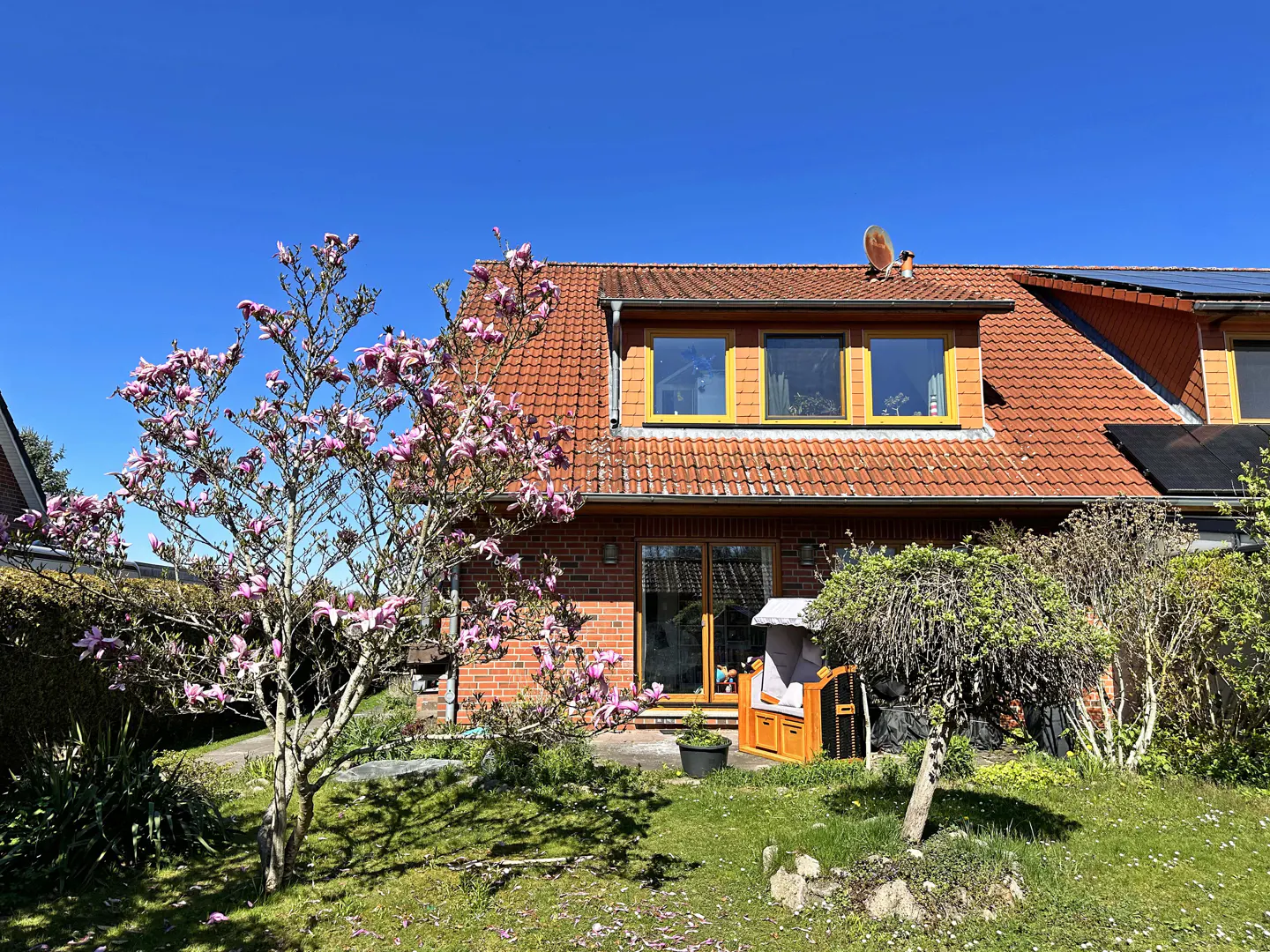 A brick house with a red tile roof, a blooming magnolia tree, and a beach chair in the yard on a sunny day.