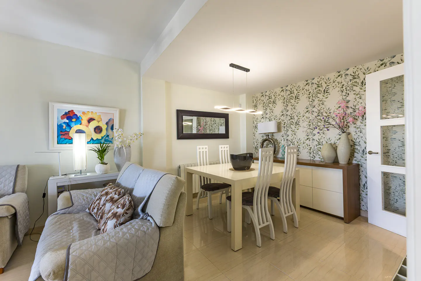 Living and dining area with a beige sofa, white table and chairs, floral wallpaper, and colorful sunflower art.
