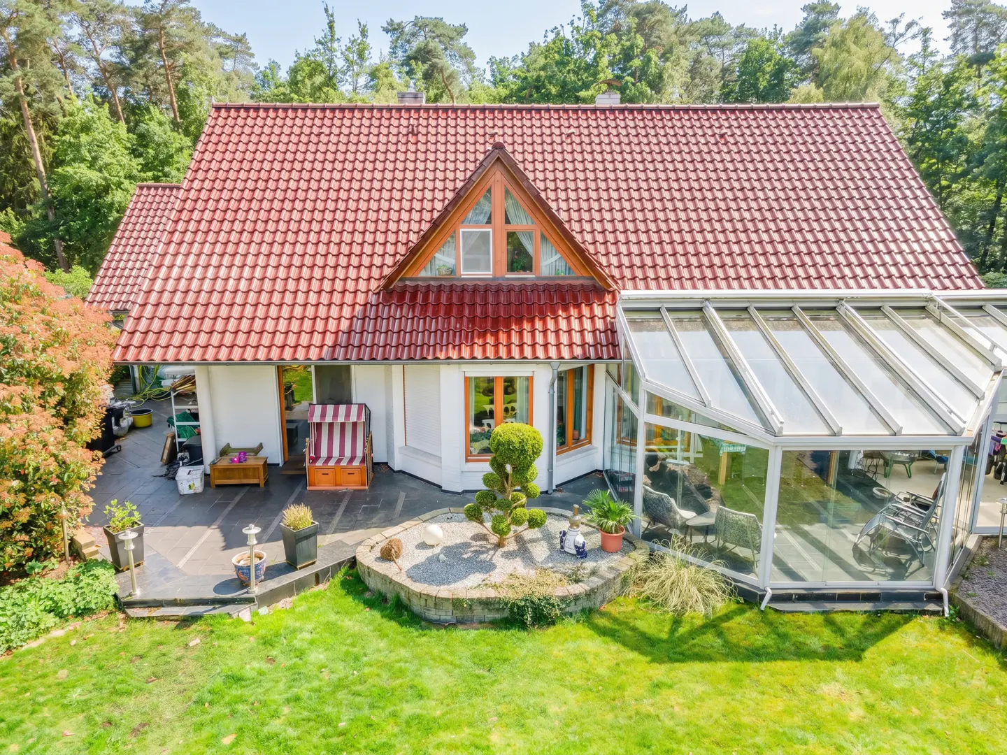 White house with a red tiled roof, a glass conservatory, and a patio area with a beach chair, set in a lush green garden.
