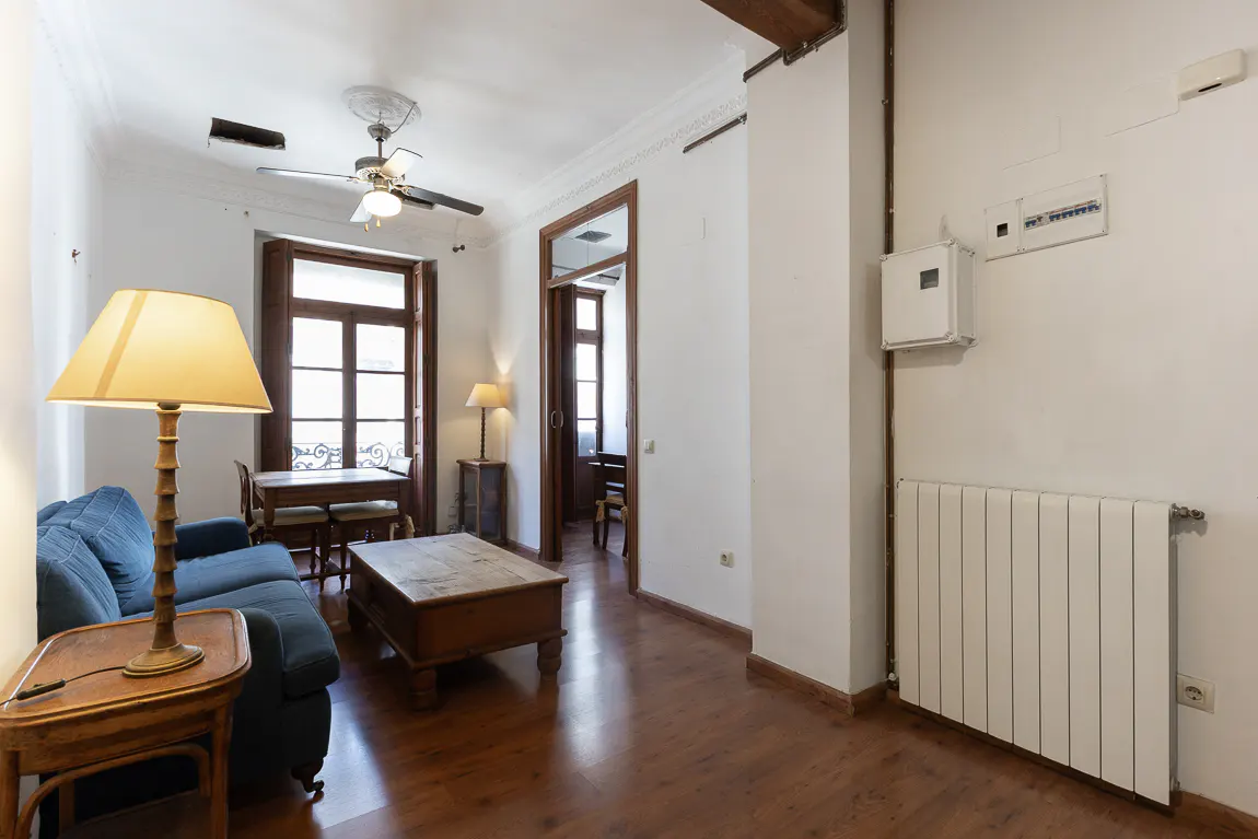 Living room with blue sofa, wood floors, and white walls. A table lamp sits on a side table next to the sofa. A ceiling fan hangs from the ceiling.