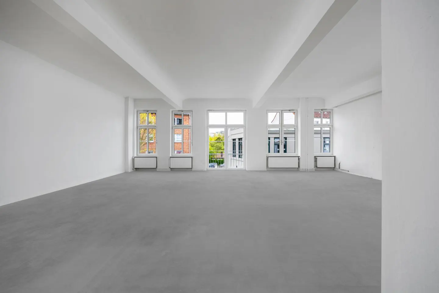 Bright, empty room with white walls, gray floor, and large windows. Radiators are under the windows. Outside view of trees and buildings.