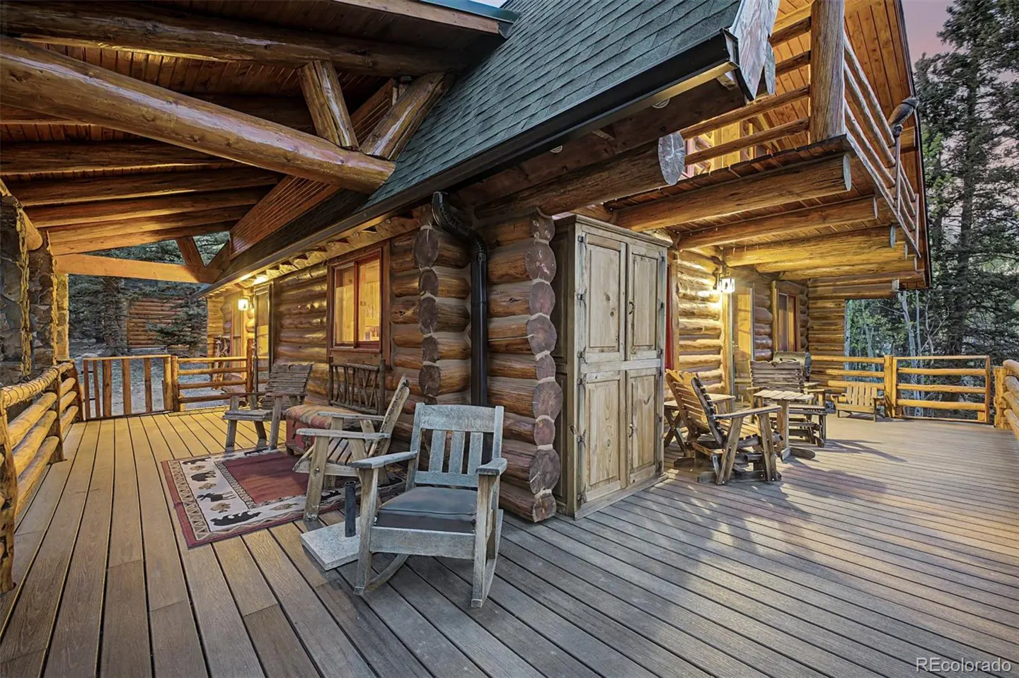 Log cabin exterior with a wide wooden porch, rocking chairs, and a bear-themed rug. The cabin has a green roof and log railings.