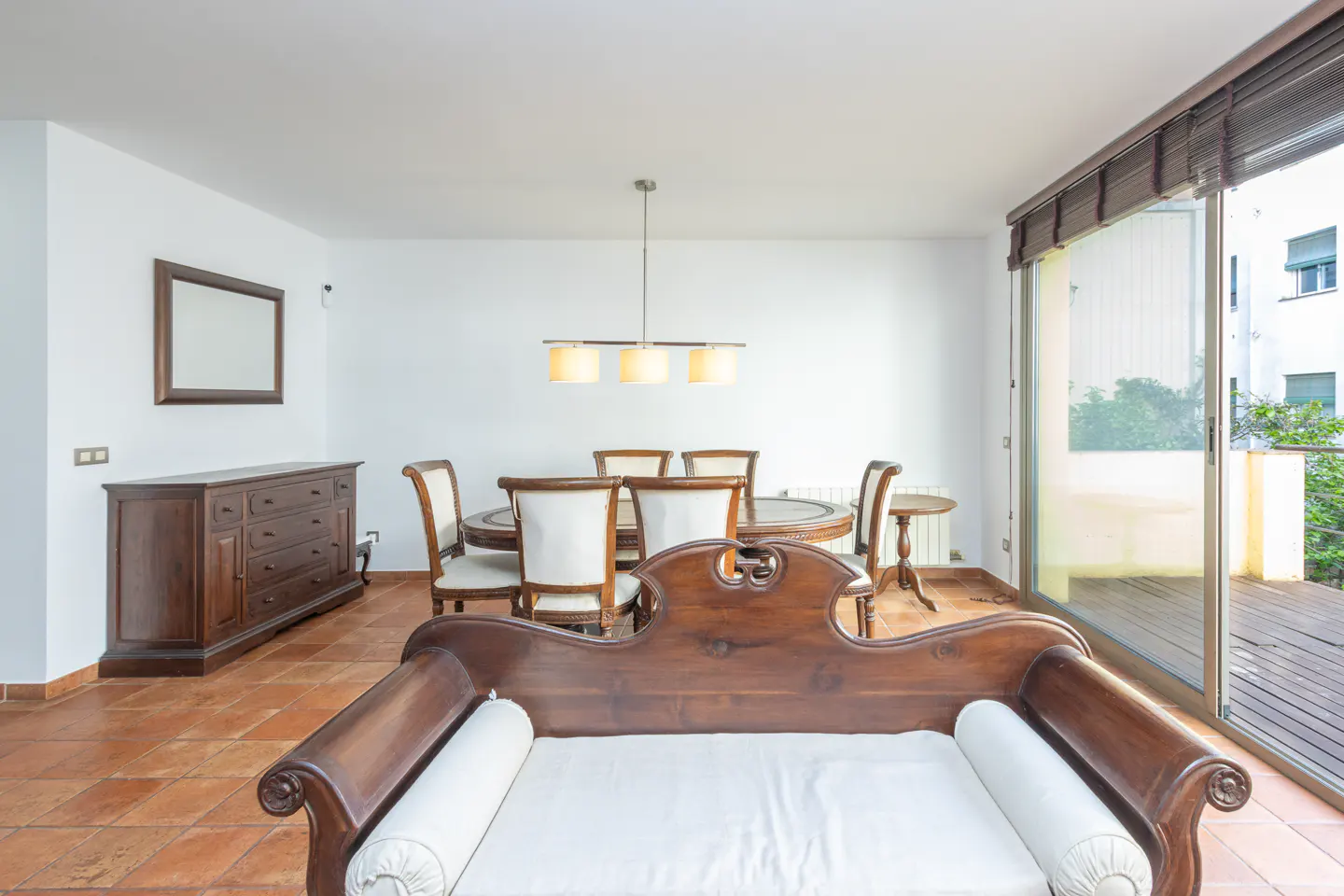 Bright dining room with white walls, terracotta tile floor, and dark wood furniture. A glass door leads to a balcony.