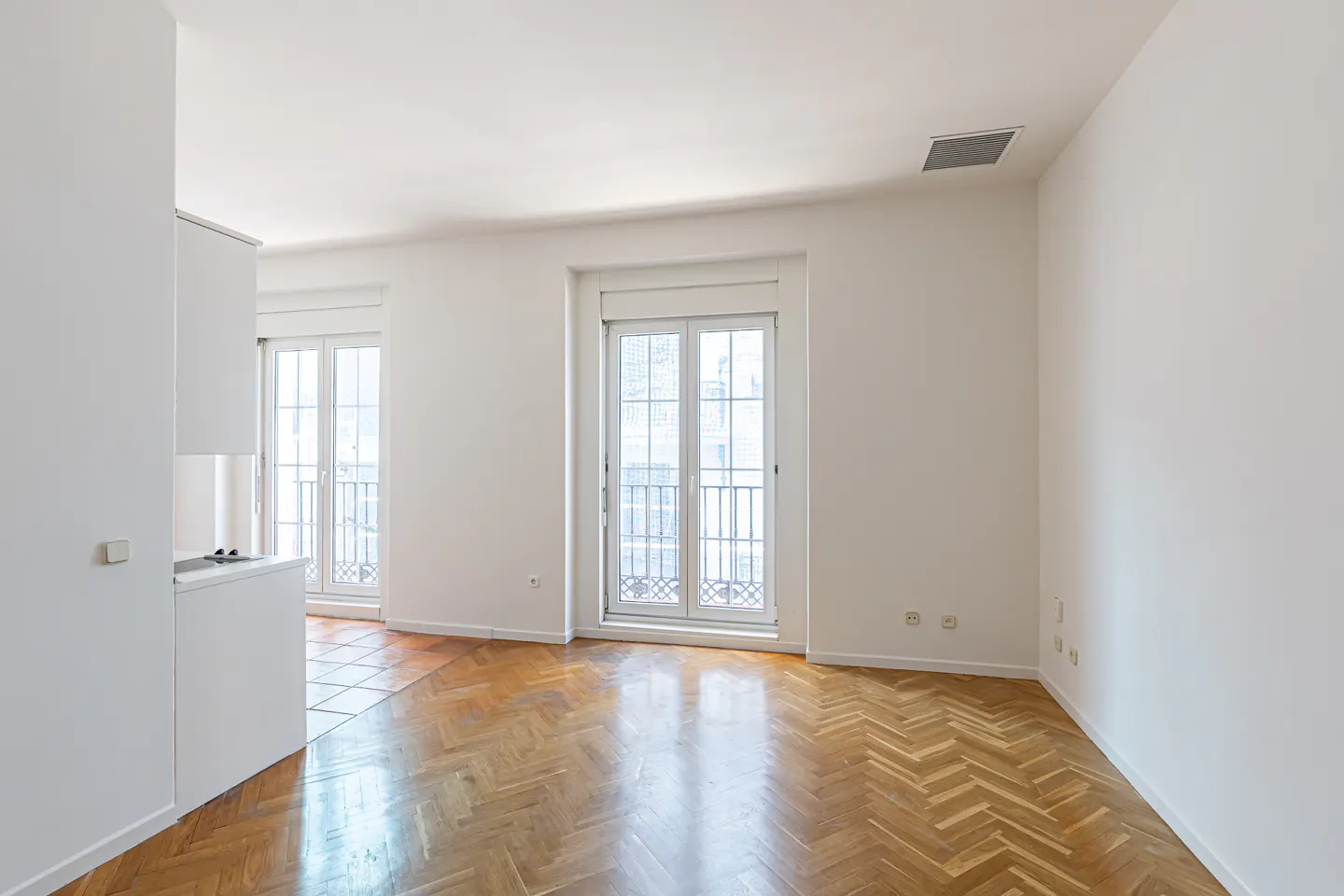 Bright, empty room with herringbone wood floors and white walls. Two sets of French doors lead to balconies.