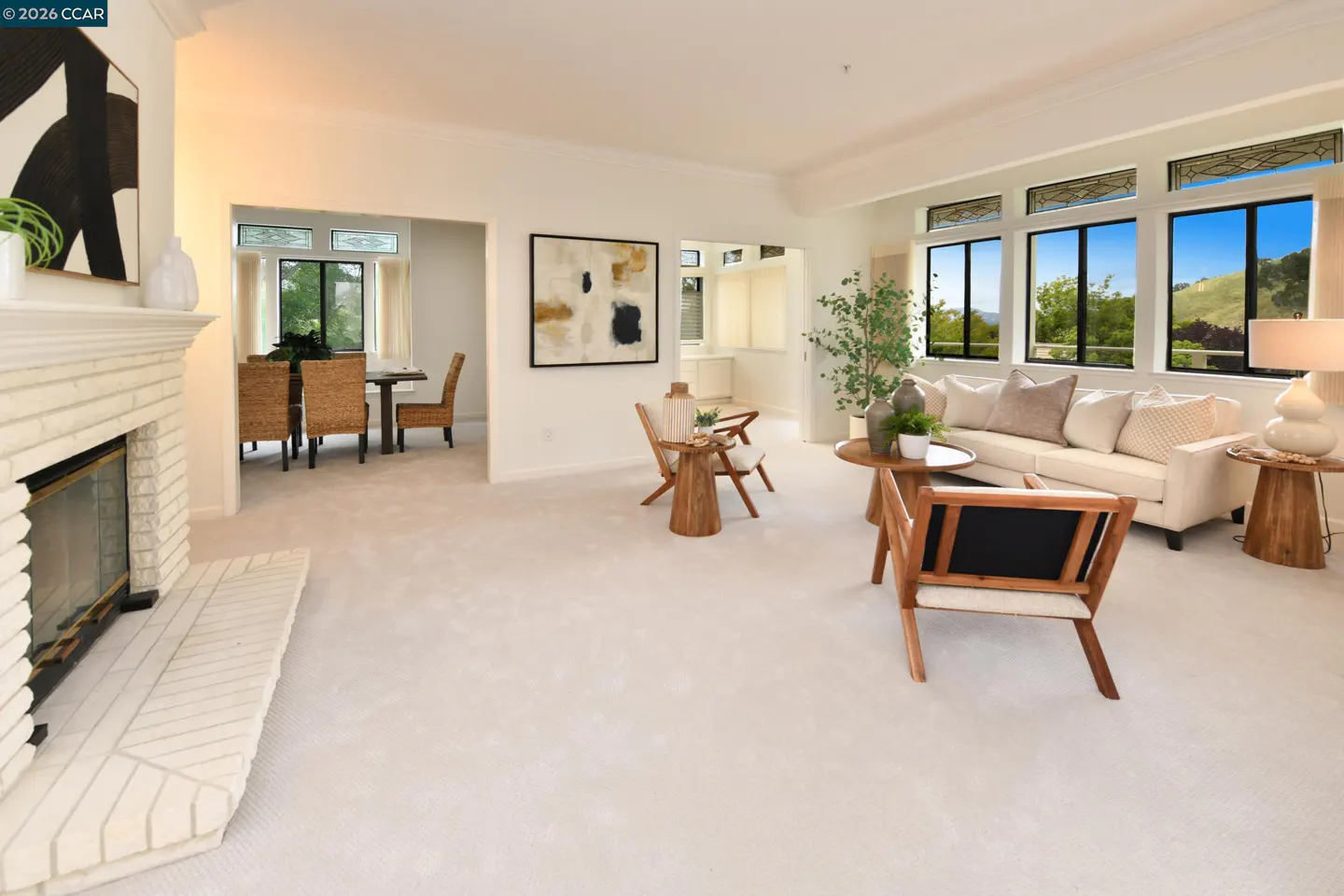 Bright, airy living room with white carpet, fireplace, sofa, and wood chairs. Large windows offer a view of green hills. Dining area visible.