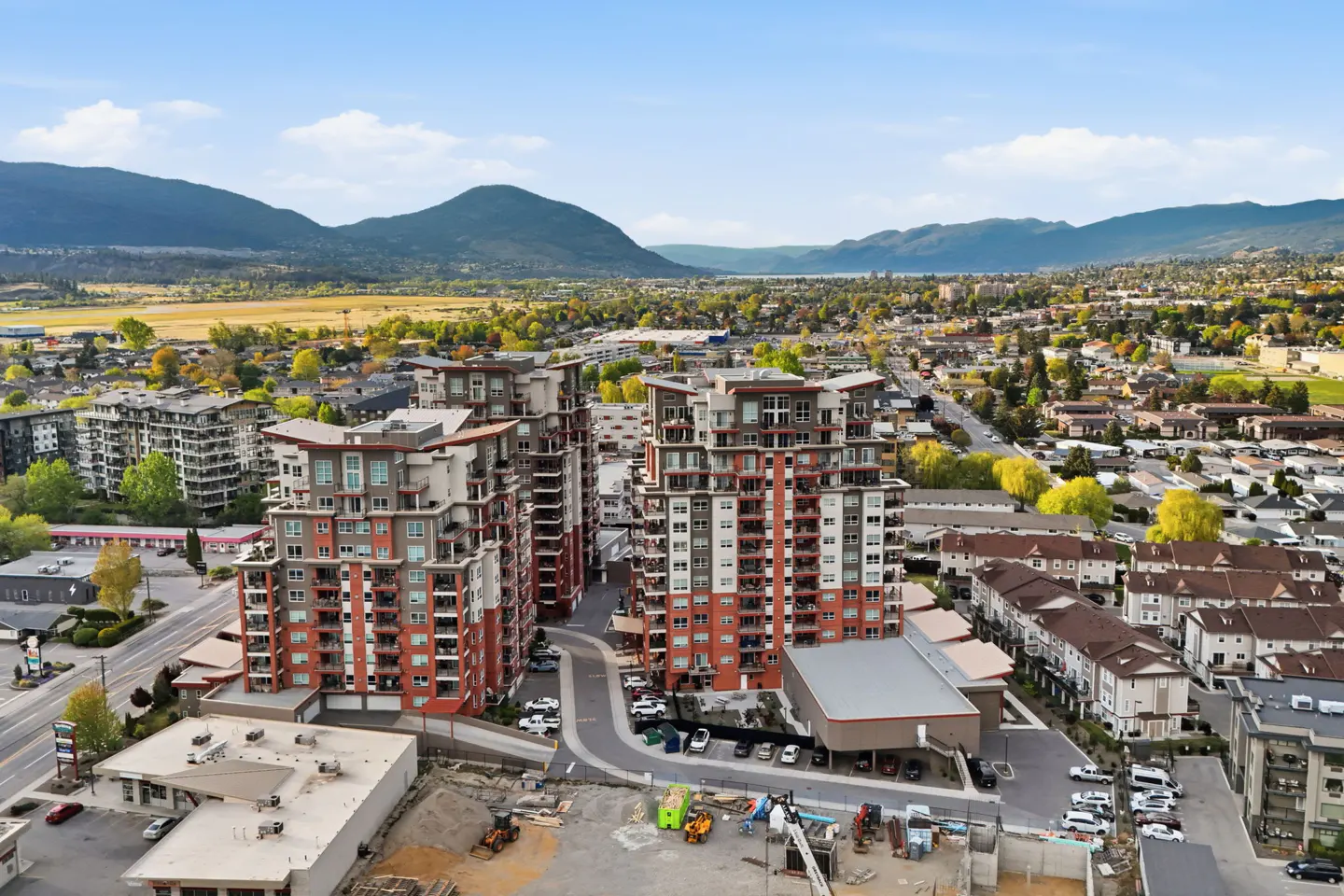 Aerial view of two modern, multi-story apartment buildings with red and gray exteriors, set against a backdrop of mountains and a city.