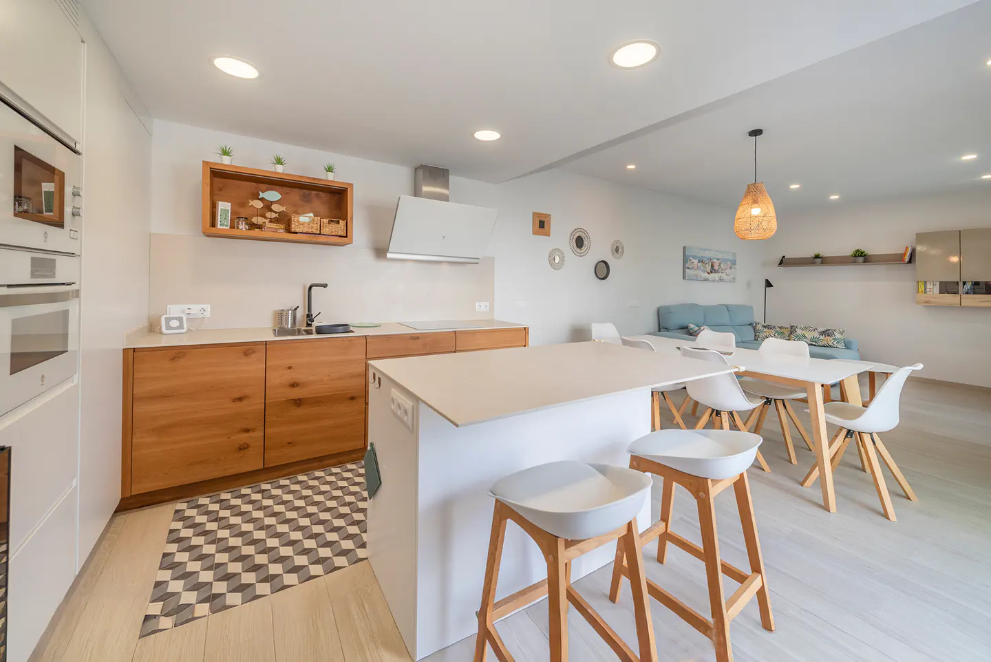 Bright, modern kitchen with wood cabinets, white island with stools, and dining area leading to a living room with a blue sofa.