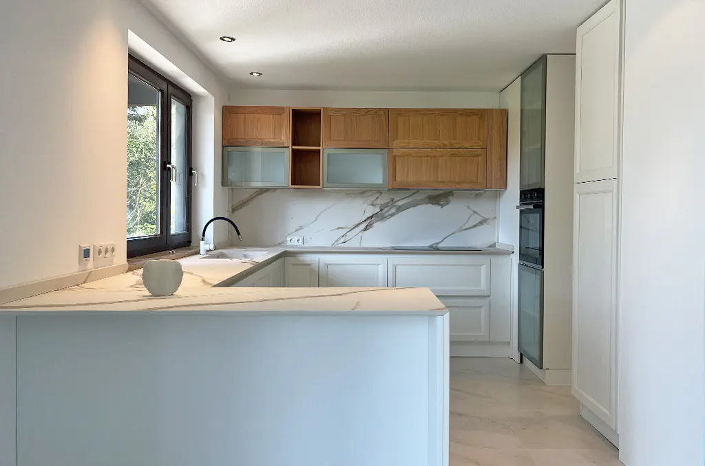 Bright kitchen with white marble counters, white cabinets, and wood cabinets above. A black faucet is visible by the window.