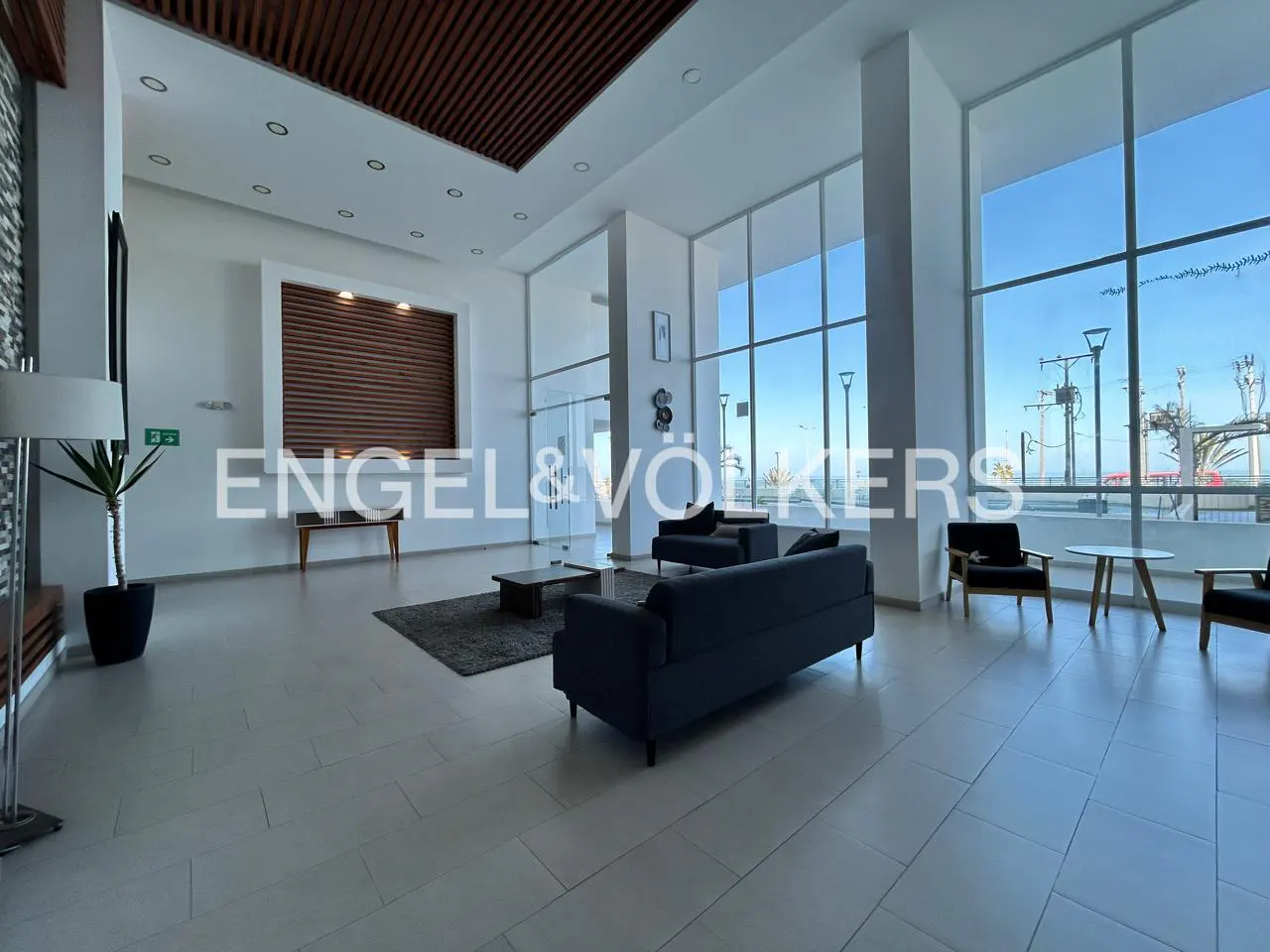 Bright, modern lobby with gray sofas, chairs, and a view of the ocean through large windows. A plant sits near a lamp.