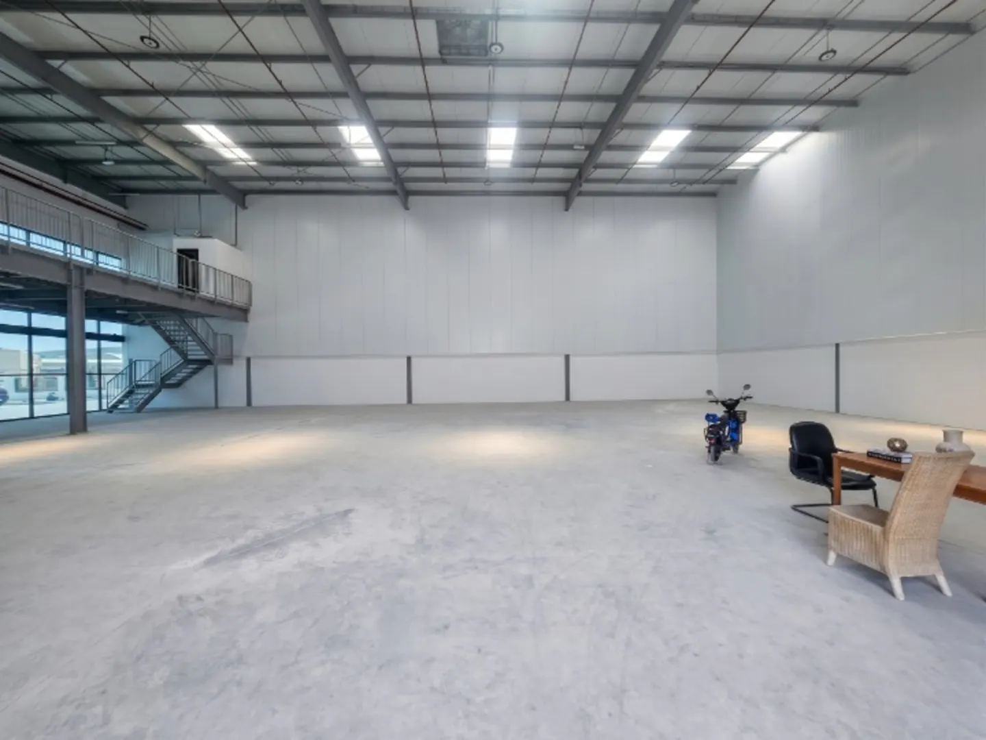 A large, empty warehouse with a concrete floor, white walls, and a metal ceiling. A blue scooter and office furniture are in the back.