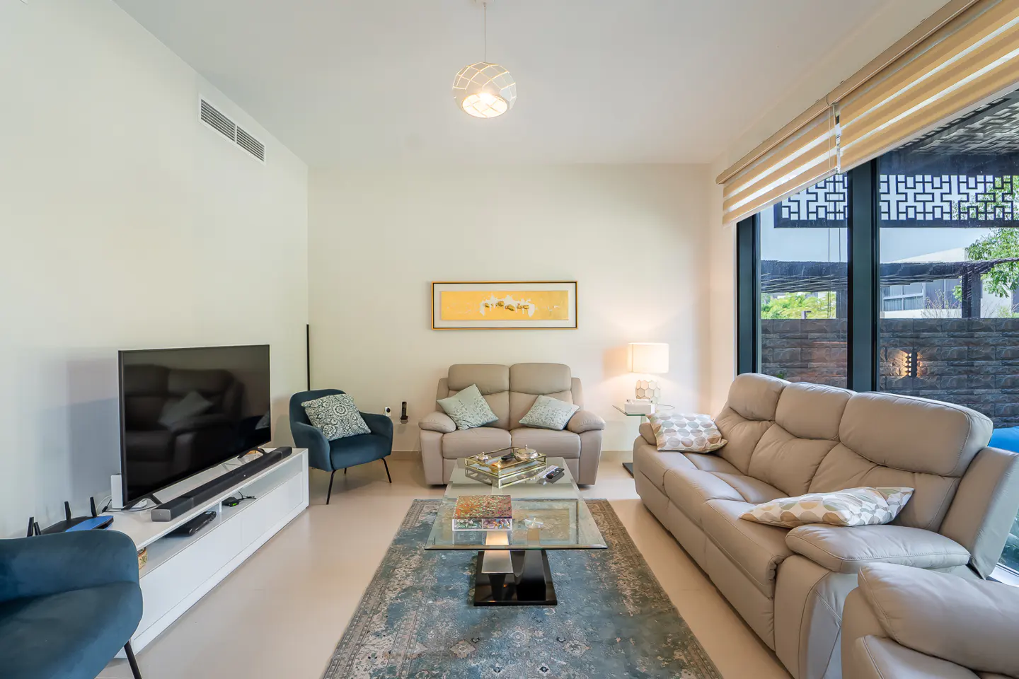 Living room with beige sofas, blue chairs, and a glass coffee table on a blue patterned rug. A large window shows an outdoor area.