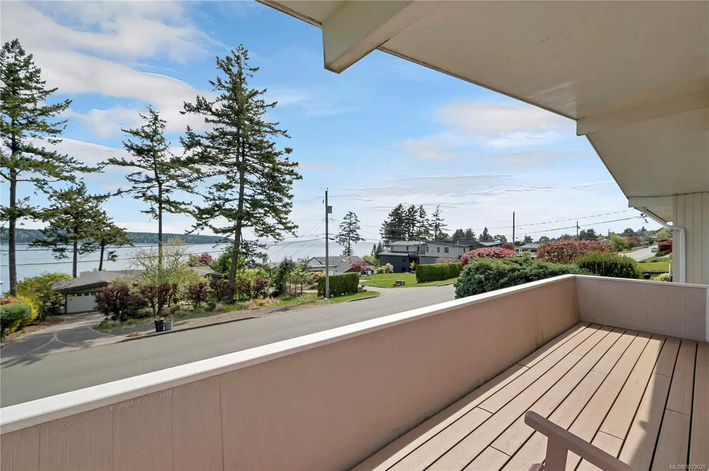View from a wood deck with a partial chair, overlooking a street, houses, trees, and a body of water under a blue sky.
