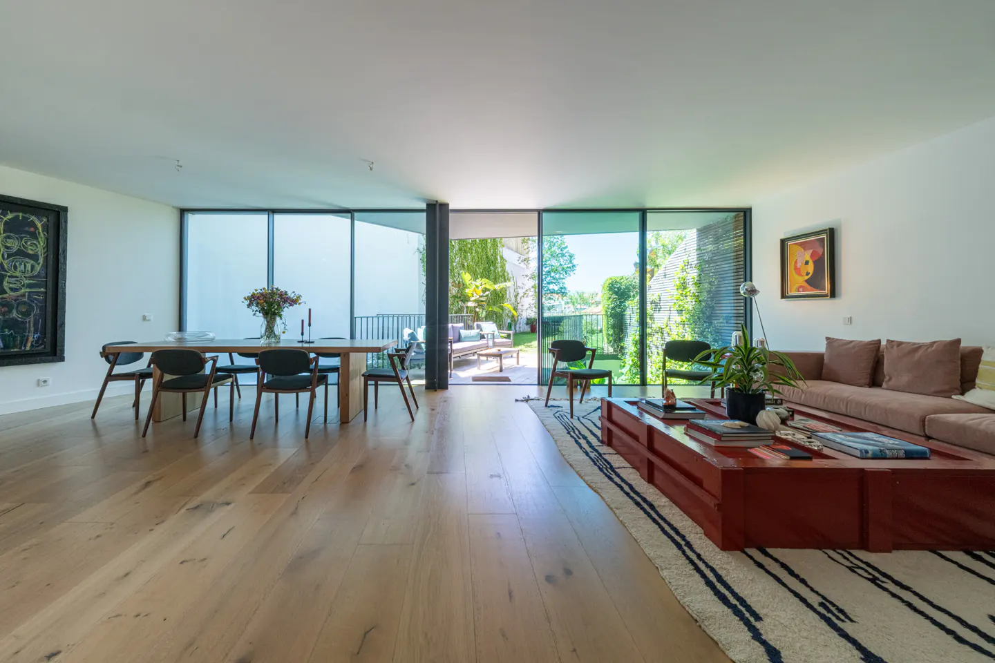 Modern open-plan living room with light wood floors, a red coffee table, a beige sofa, and floor-to-ceiling glass doors.