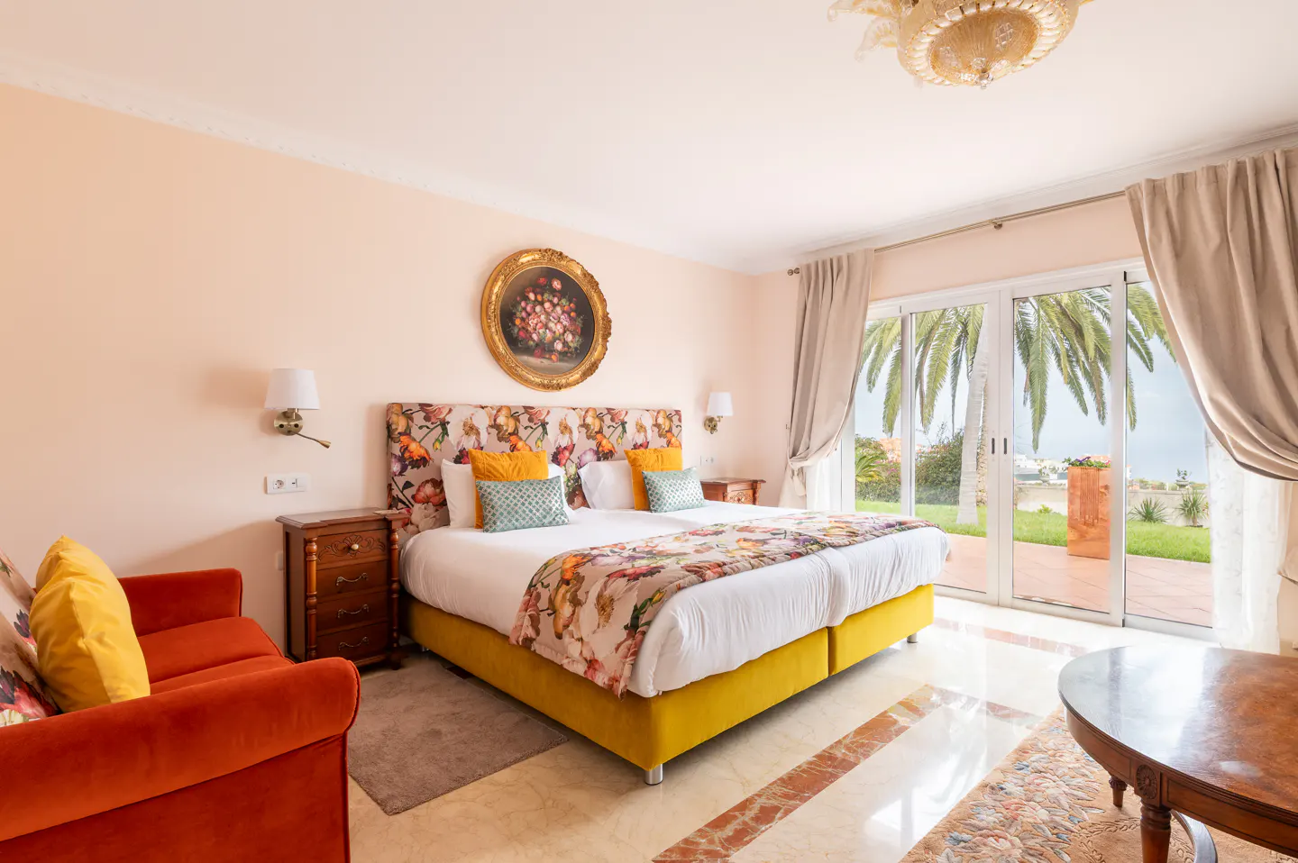 Bright bedroom with a floral headboard, yellow bed frame, and orange sofa. Sliding glass doors lead to a patio with palm trees.