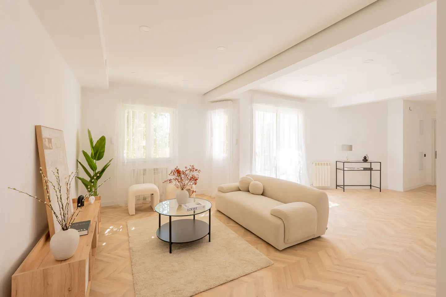 Bright, modern living room with white walls, light wood floors, and neutral furniture. A beige sofa, glass coffee table, and plants add warmth.