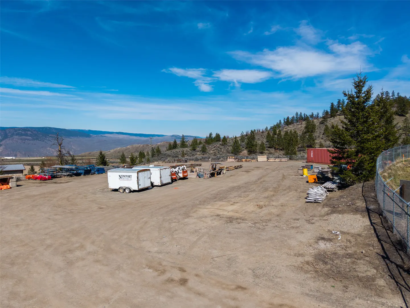 Wide shot of a dirt lot with trailers, equipment, and a red shipping container under a blue sky. Hills and trees in the background.