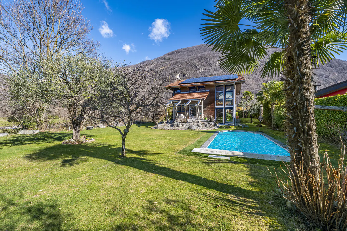 A modern wood home with a pool sits on a green lawn with trees and a mountain backdrop.