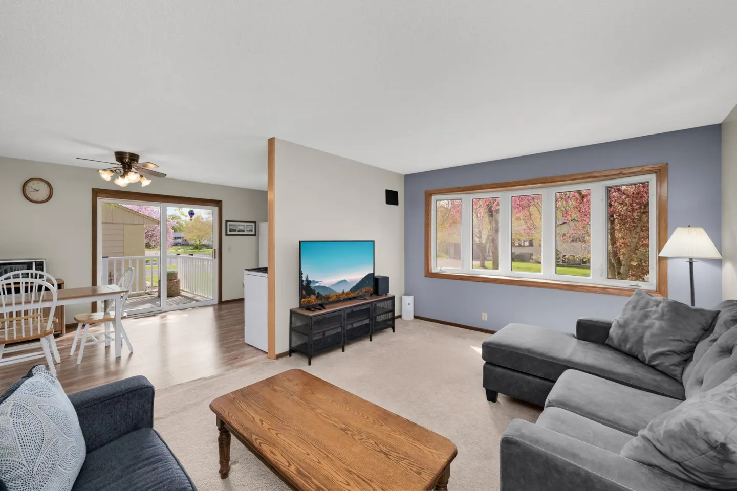 Living room with gray sofa, wood coffee table, and TV. A dining area is visible through a doorway. Outside, trees with pink blossoms are in bloom.