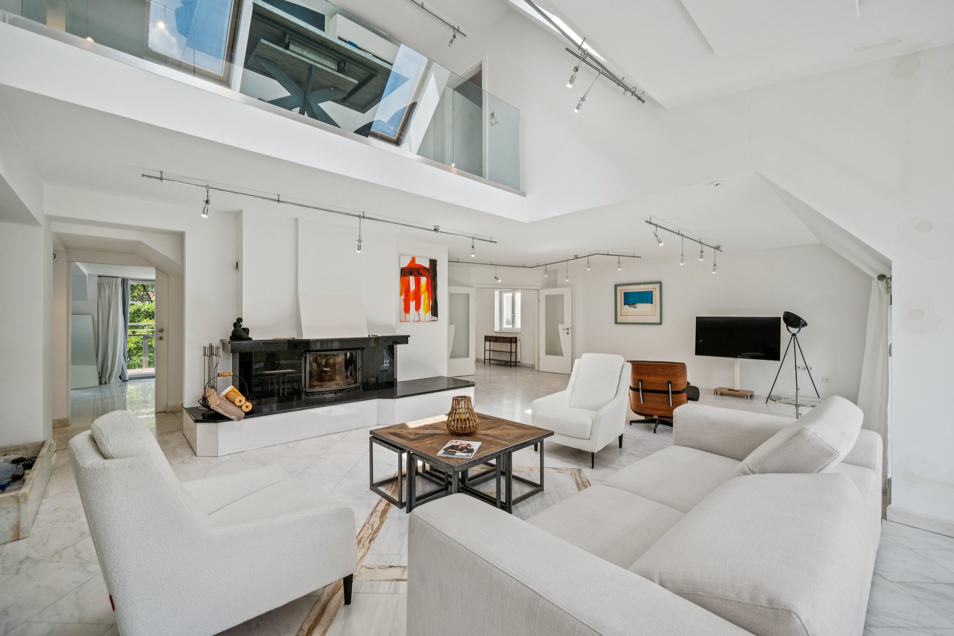 Spacious, modern white living room with a fireplace, white sofas, armchairs, a wooden coffee table, and a mezzanine level.