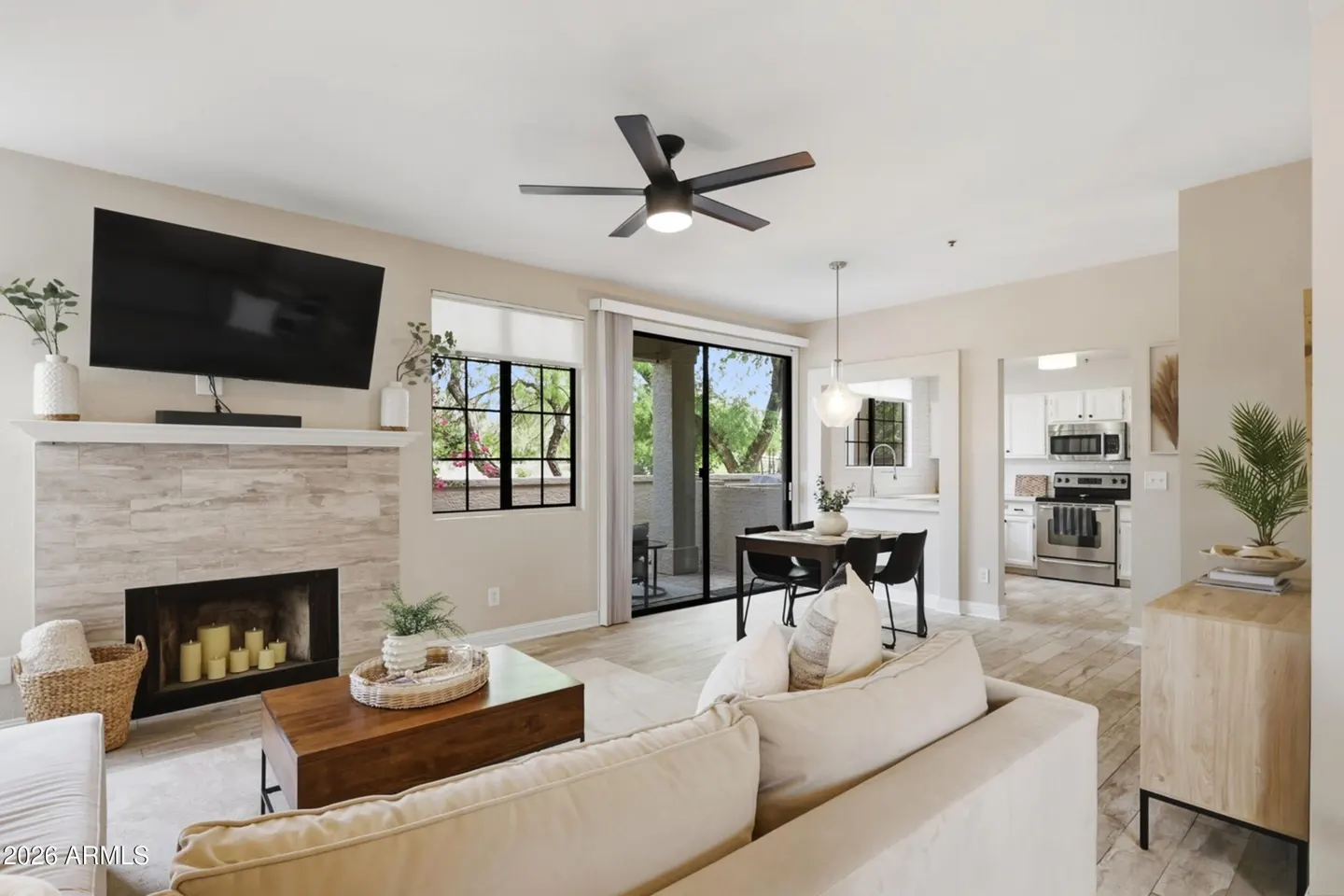 Bright living room with a fireplace, TV, and a cream-colored sofa. A dining area and kitchen are visible in the background.