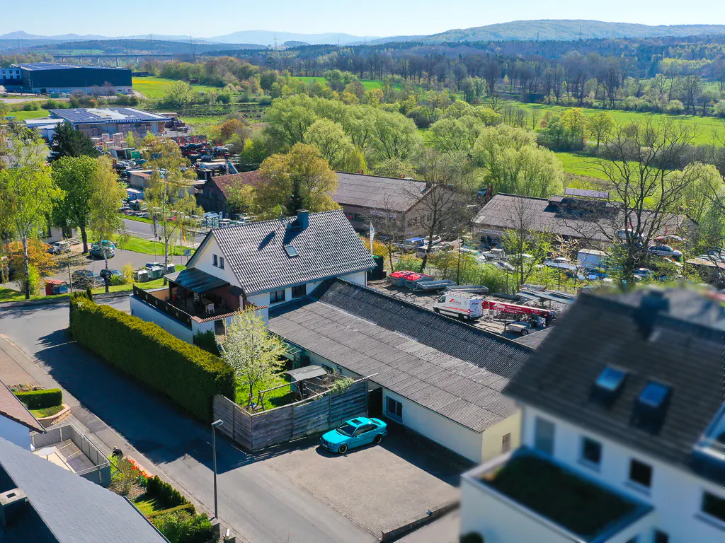 Aerial view of a house with a gray roof, a blue car parked in the driveway, and green trees in the background.