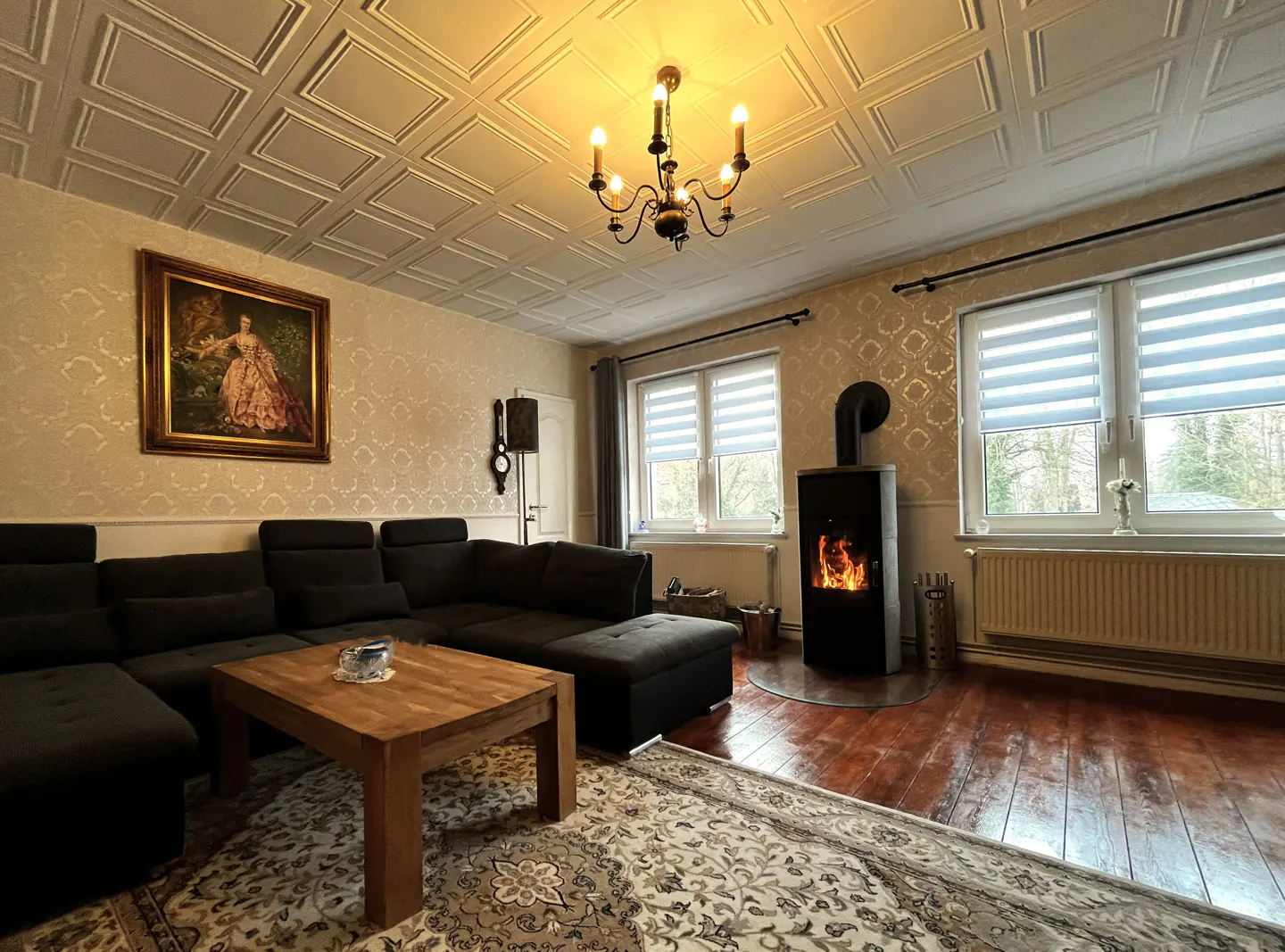 Living room with a black sectional sofa, wood coffee table, and a black fireplace with a visible fire. The room has a patterned rug and wallpaper.