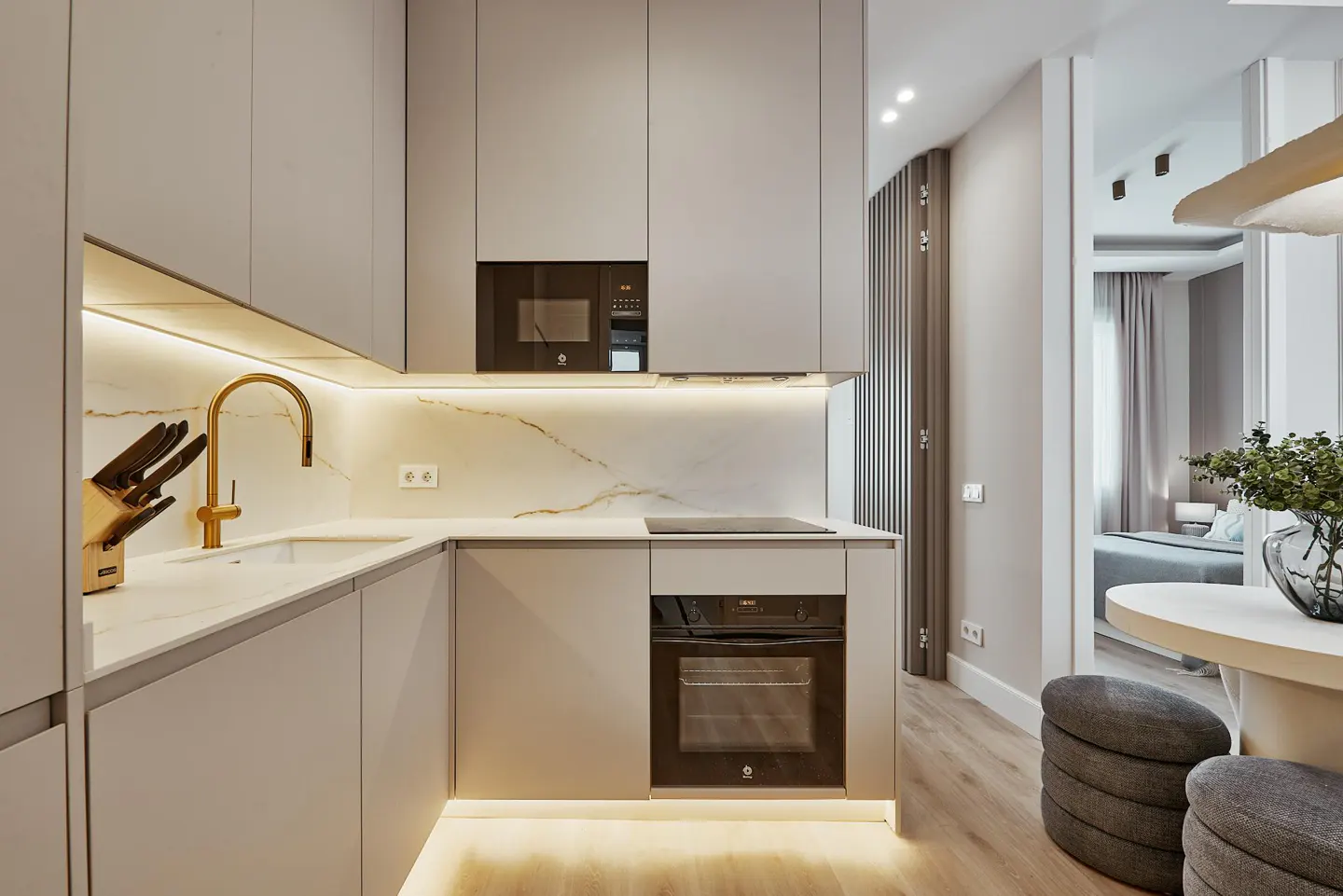 Modern kitchen with light gray cabinets, gold faucet, marble backsplash, and black appliances. View into dining area and bedroom.