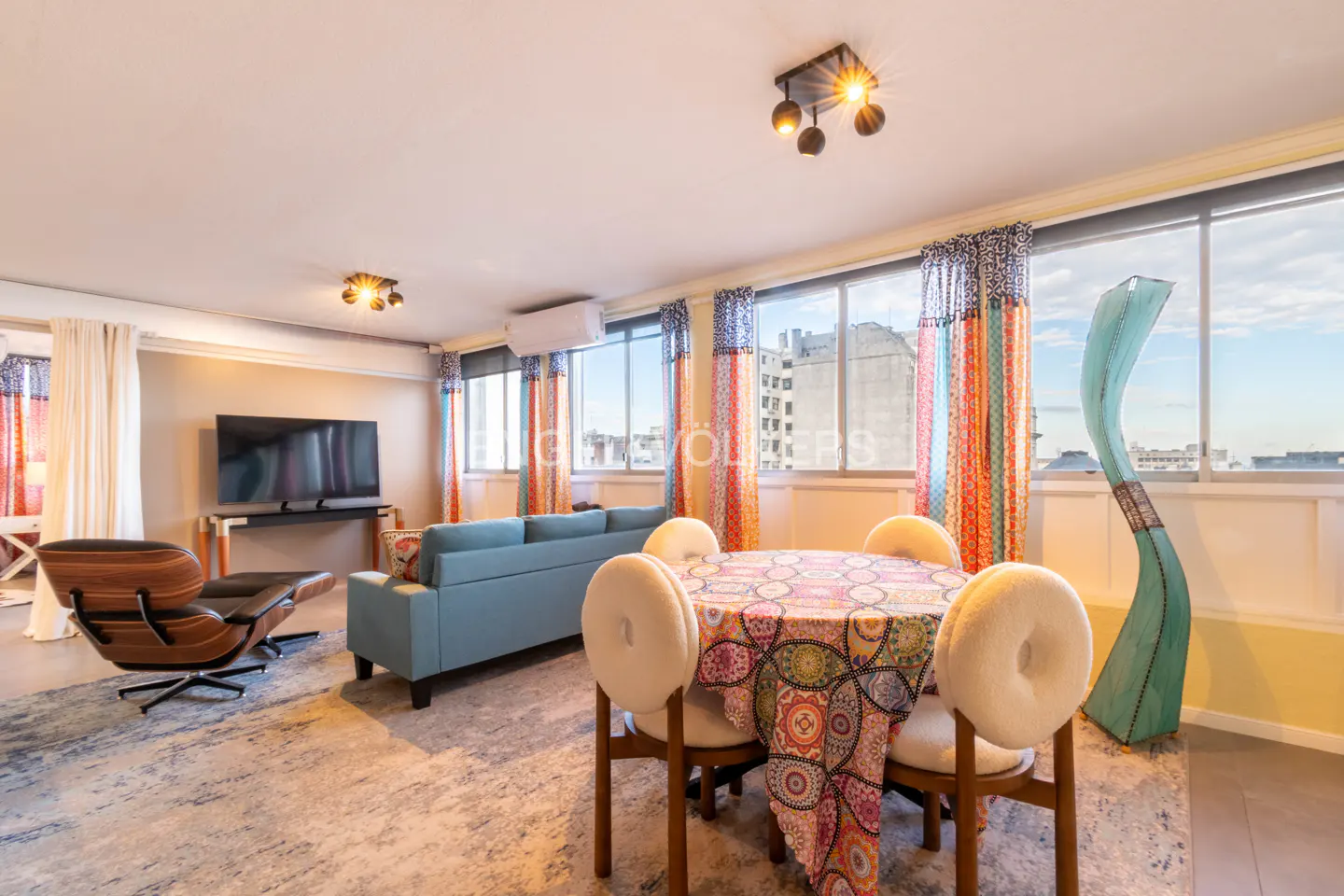 Bright living room with a blue sofa, round dining table with colorful tablecloth, and a modern chair. Large windows offer a city view.