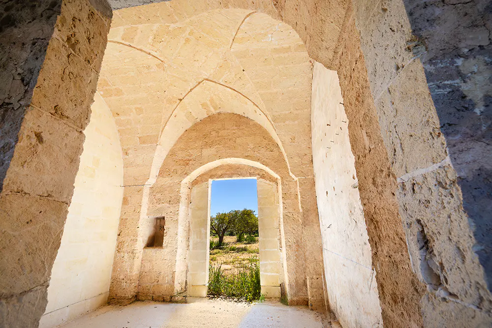 Interior view of a stone building with arched ceilings, looking out to a field with a tree under a blue sky.