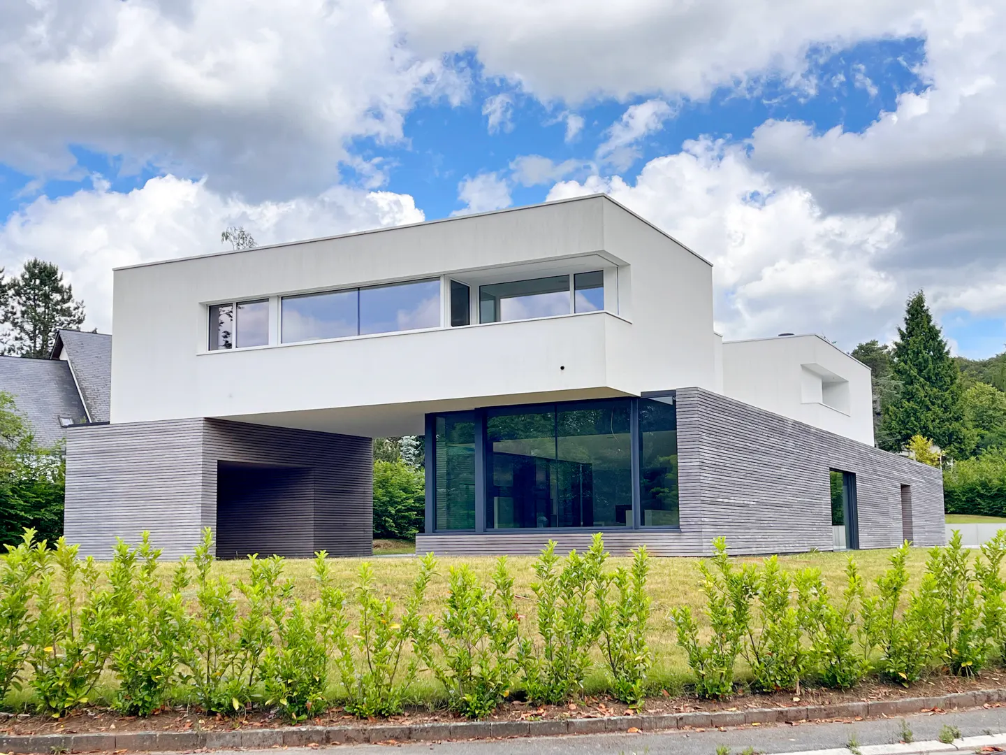 Modern white and grey horizontal home with large glass windows, set behind a green hedge under a cloudy blue sky.