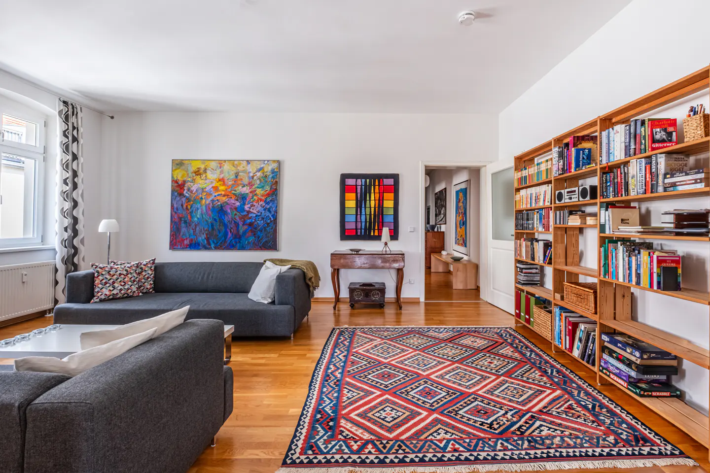 A bright living room with hardwood floors, gray sofas, colorful art, and a large wooden bookshelf.