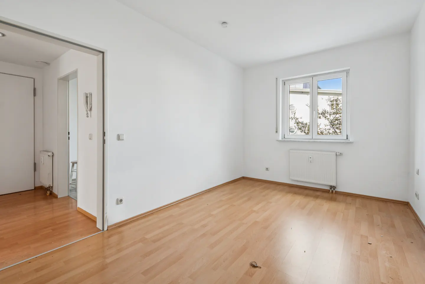 Bright, empty room with wood floor, white walls, and a window. A doorway leads to another room with a white door.