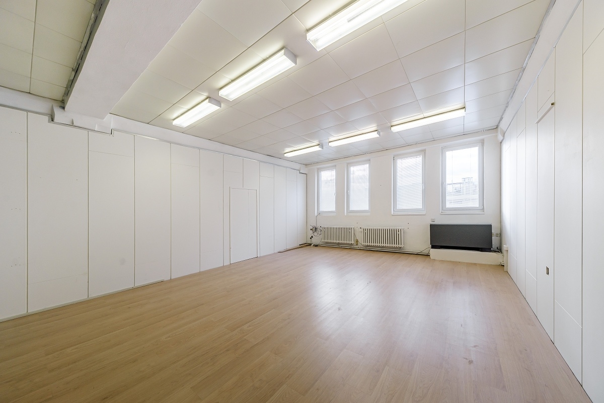Bright, empty room with light wood floors, white walls, and fluorescent lighting. Windows line the back wall. Radiators are visible. [AI-generated text]